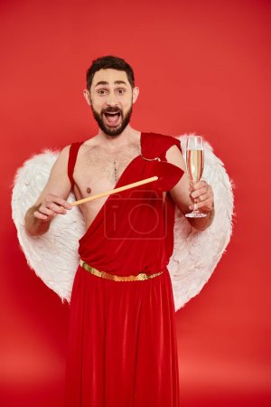 thrilled bearded cupid man with heart-shaped arrow and champagne glass looking at camera on red