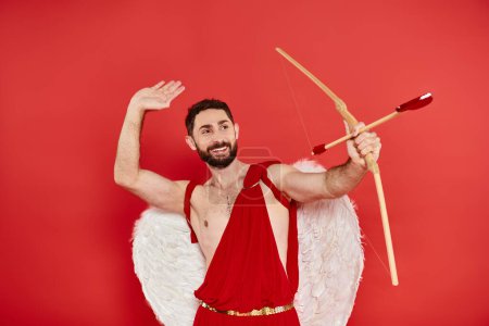 cheerful man in cupid costume archering with heart-shaped arrow on red, st valentines costume party