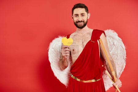 hombre barbudo alegre en traje de cupido con arco y corazón de papel amarillo en rojo, día de San Valentín