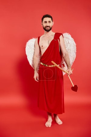 smiling barefoot bearded man in cupid costume with bow and arrow standing and looking away on red