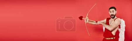 thrilled cupid man with open mouth archering while looking at camera on red, horizontal banner