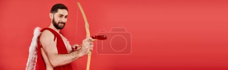 joyful bearded man dressed as cupid shooting from bow with heart-shaped arrow on red, banner