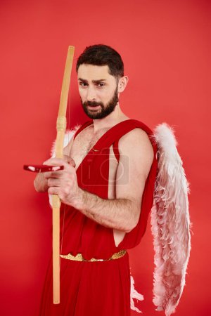 serious man in cupid costume with bow and heart-shaped arrow looking at camera on red backdrop