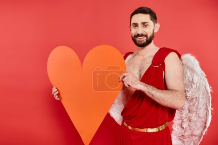 pleased bearded man in cupid costume holding huge orange paper heart on red, Saint Valentines day