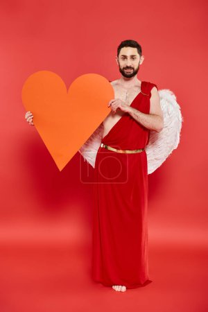 full length of bearded man dressed as cupid with huge paper heart on red, valentines costume party