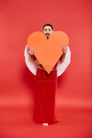 smiling bearded man dressed as cupid holding huge orange heart on red, valentines costume party