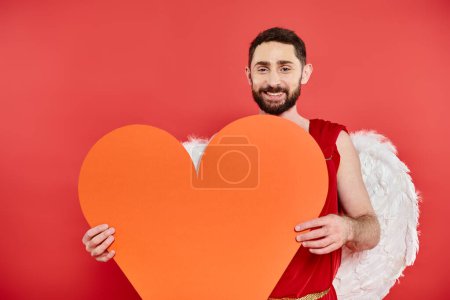 freudiger bärtiger Mann mit Amor-Flügeln, der am Valentinstag ein riesiges orangefarbenes Herz auf rotem Grund hält