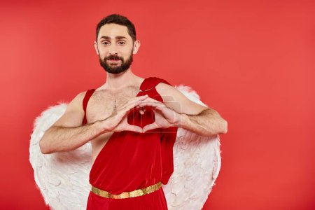 bearded man in costume and wings of cupid showing heart sign with hands and smiling at camera on red