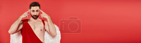 pensive bearded man in cupid costume touching head looking at camera on red, horizontal banner