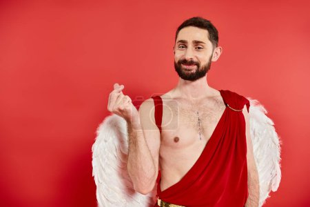 joyful bearded cupid man showing small heart sign with fingers and looking at camera on red