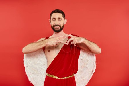 pleased bearded cupid man showing heart sign with fingers and smiling at camera on red backdrop