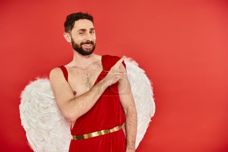 Téléchargez les photos : Joyeux homme barbu Cupidon regardant la caméra et pointant du doigt sur rouge, st Saint-Valentin - en image libre de droit