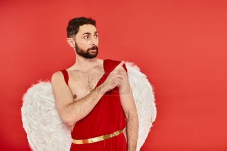 Téléchargez les photos : Homme sérieux en costume de Cupidon regardant loin et pointant du doigt sur rouge, st Saint-Valentin - en image libre de droit