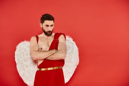serious and thoughtful cupid man with folded arms standing on red backdrop, Saint Valentines concept