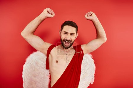 excited bearded cupid man rejoicing and showing win gesture with raised hands on red backdrop