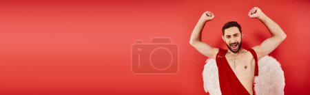 Téléchargez les photos : Joyeux homme barbu en costume de Cupidon montrant geste de victoire avec les mains levées sur fond rouge, bannière - en image libre de droit