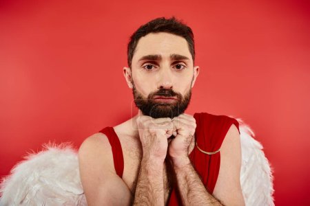 portrait of frustrated bearded man dressed as cupid looking at camera on red, st valentines concept