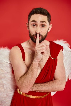 funny bearded man dressed as cupid showing hush sign and looking at camera on red, st valentines day