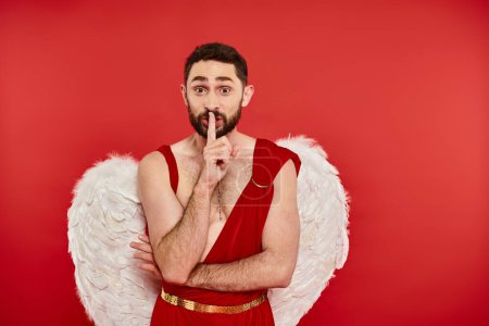 excited bearded man in cupid costume showing shh sign and looking at camera on red, st valentines