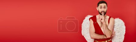 bearded man dresses as cupid looking at camera and showing hush sign on red, horizontal banner