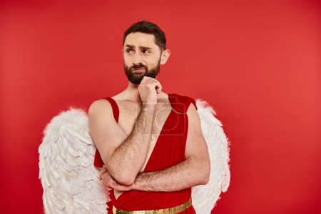 displeased bearded man in cupid costume holding hand near chin and  looking away on red backdrop