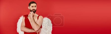 Téléchargez les photos : Homme barbu insatisfait en costume de Cupidon tenant la main près du menton et regardant loin sur rouge, bannière - en image libre de droit
