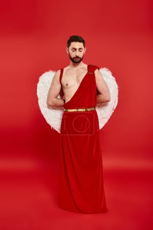 full length of thoughtful bearded man in cupid costume standing with hands behind back on red