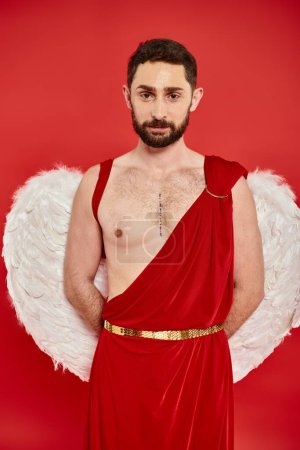 bearded man in cupid costume and wings posing with hands behind back and looking at camera on red