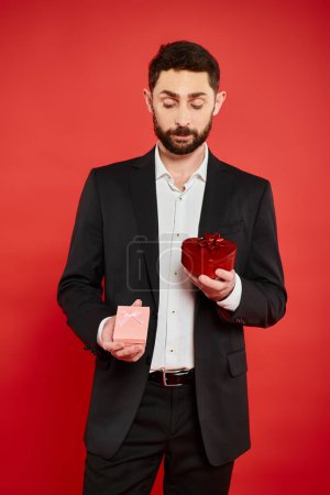 thoughtful bearded man in black formal wear choosing Saint Valentines day present on red backdrop