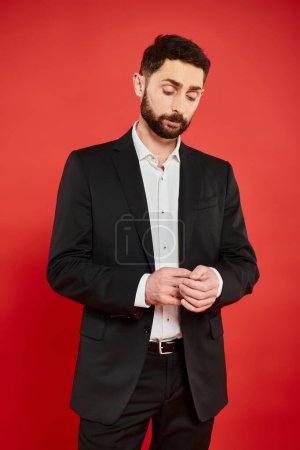 Nachdenklicher bärtiger Geschäftsmann im schwarzen eleganten Anzug steht und denkt auf rotem Hintergrund im Studio