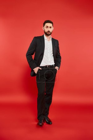 selbstbewusster und stilvoller Geschäftsmann im schwarzen Anzug mit Händen in Taschen auf rotem, durchgehendem Stoff
