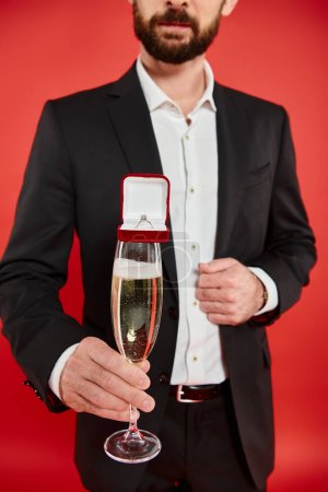cropped view of man in black suit with champagne glass and gift box with jewelry ring on red
