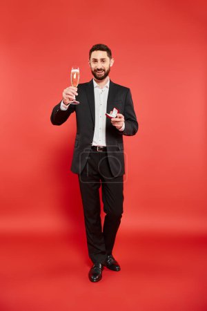 happy elegant man in black suit with champagne glass and jewelry box on red, st valentines day