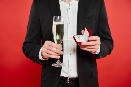 cropped view of elegant man with champagne glass and ring in jewelry box on red, st valentines gift