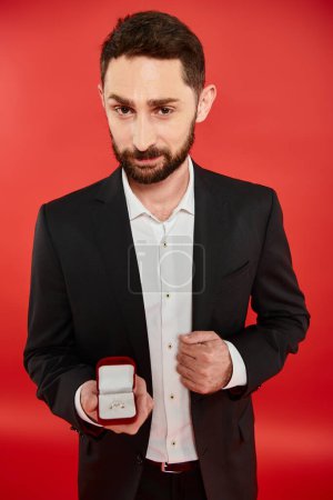 confident bearded man in black suit holding jewelry box on red, present on st valentines day