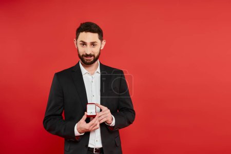 smiling bearded man in black suit showing ring in jewelry box on red, st valentines present