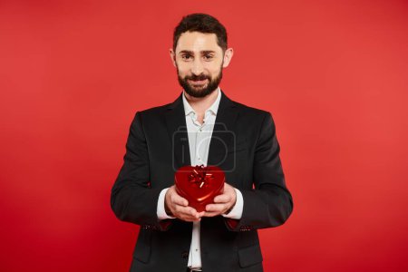hombre barbudo alegre en traje elegante que muestra la caja de regalo en forma de corazón en rojo, día de San Valentín