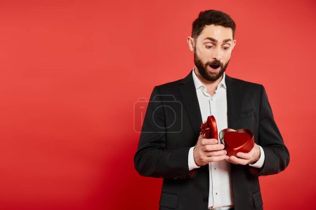 amazed man in black suit opening heart-shaped gift box on red, Saint Valentines day present