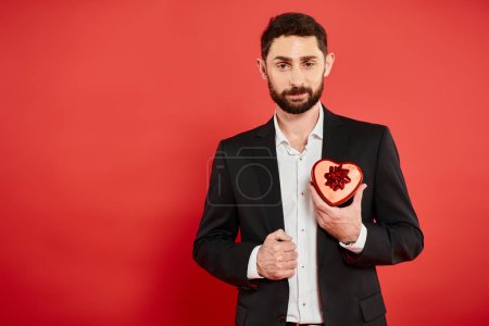 pleased bearded businessman in black suit with heart-shaped gift box on red, Saint Valentines day