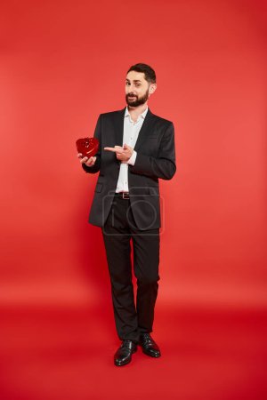 full length of elegant man in black suit pointing at heart-shaped gift box on red, st valentines day