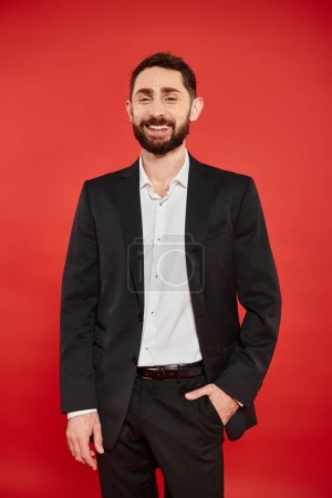 cheerful bearded businessman in black suit posing with hand in pocket and laughing on red backdrop