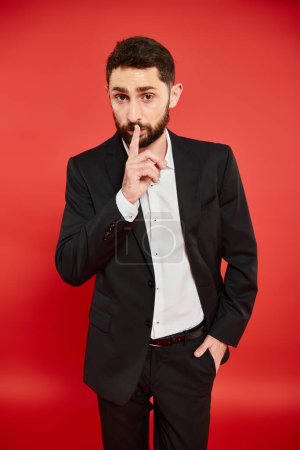 bearded man in black suit showing hush sign while standing with hand in pocket on red backdrop