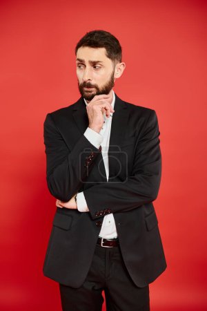 pensive businessman in black suit thinking and looking away while standing on red backdrop