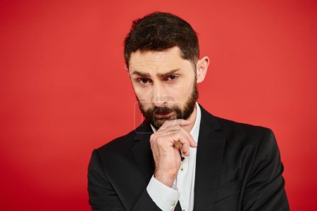 serious bearded businessman in black suit with hand near chin looking at camera on red backdrop