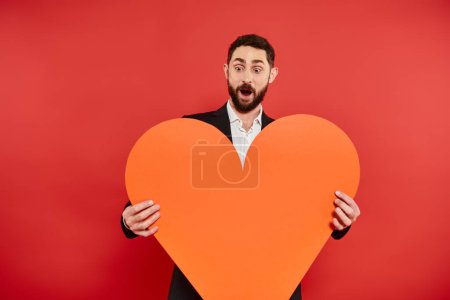 amazed bearded man with open mouth and huge orange paper heart on red, Saint Valentines concept
