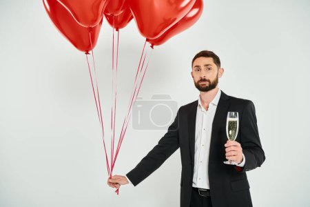 elegante hombre con globos rojos en forma de corazón y copa de champán en gris, concepto de San Valentín