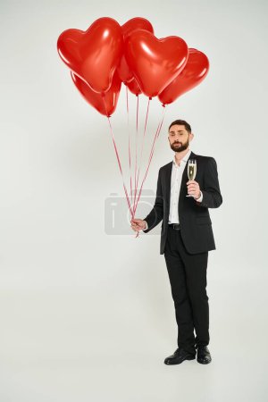 Photo for Wealthy man in black suit with champagne and red heart-shaped balloons on grey, st valentines day - Royalty Free Image