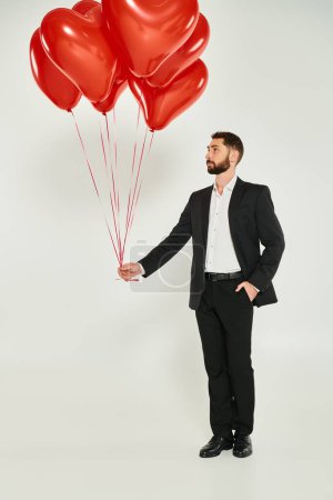Photo for Full length of man in black elegant suit with red heart-shaped balloons and hand in pocket on grey - Royalty Free Image