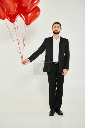 hombre de negocios barbudo en traje negro con globos rojos en forma de corazón mirando a la cámara en gris
