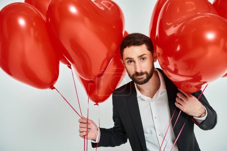 charismatic man in black suit with st valentines heart-shaped balloons looking at camera on grey